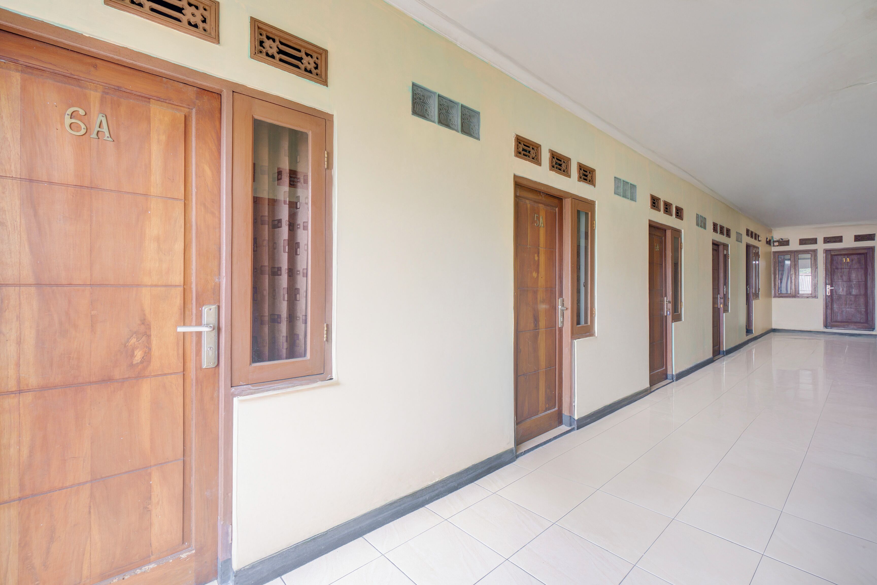 Un largo pasillo vacío con puertas de madera numeradas, piso de baldosas y pared lisa con detalles decorativos.
