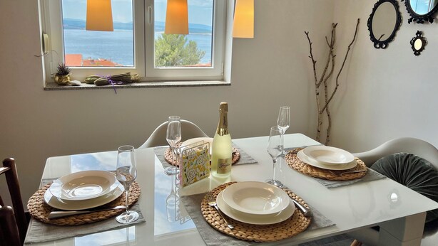 A dining table set with plates, glasses, and a bottle of champagne, with a view of the sea through a window. - Apartman Jasmin 1 (Crikvenica)