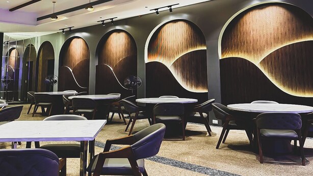 A modern restaurant interior with round tables and chairs, a patterned carpet, and a wall with wooden panels and lighting.