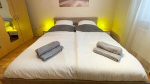 Un lit double avec une literie blanche, deux oreillers et deux serviettes pliées. Un tableau est accroché au mur, au-dessus du lit.