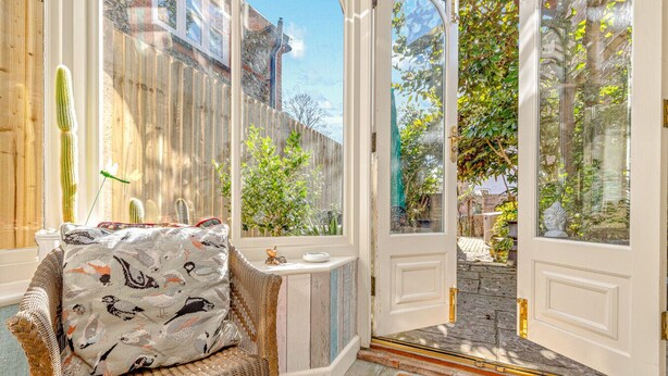 A wicker chair with a patterned cushion, a white door with a doormat, and a view of a garden through the door. - Basketmakers Cottage (Henley-On-Thames)