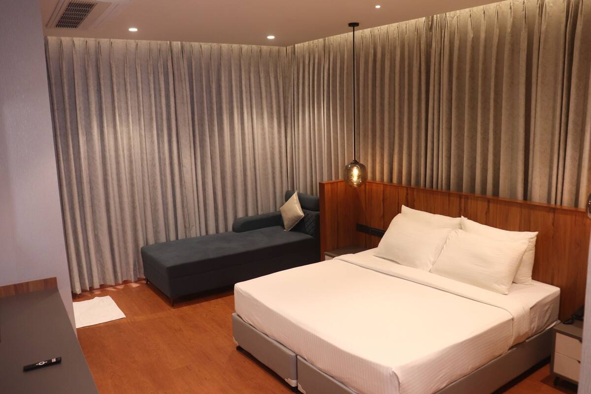 A modern hotel room with a large bed, a sofa, wooden headboard, and a curtain-covered window.
