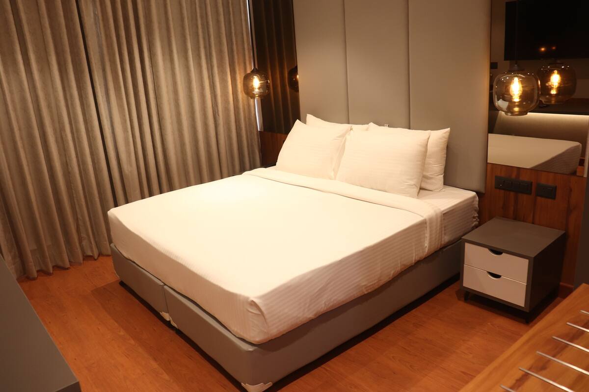 A neatly made bed with white linens, a wooden nightstand with a drawer, and a large window with curtains.