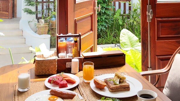 A breakfast table with eggs, toast, fruit, and beverages, set in a room with wooden doors and a view of a courtyard. - Asoca Hotel Luang Prabang (Luang Prabang)