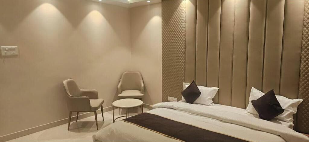 Quarto de hotel com cama, duas cadeiras, uma mesinha e uma parede com painéis verticais.