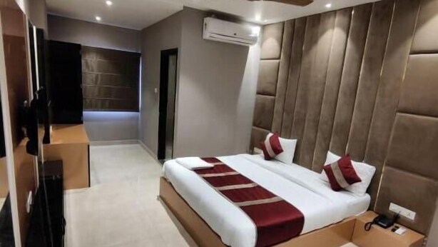 A modern hotel room with a large bed, a ceiling fan, an air conditioning unit, and a small bedside table with a phone. - Hotel Maya Crystal (Kanpur)