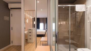 A modern hotel room with a glass shower enclosure, a wooden floor, a bed, a desk, and a wardrobe.