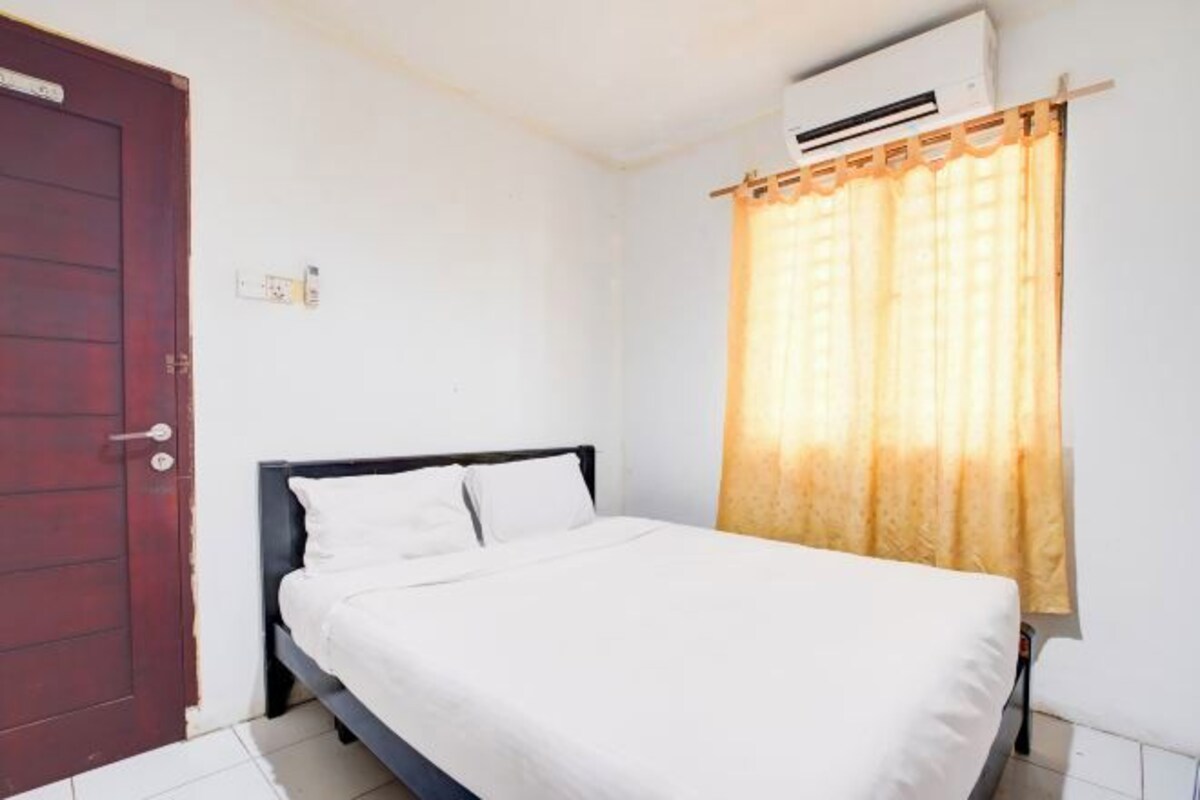 Una cama individual con ropa de cama blanca en una habitación con paredes blancas, una puerta de madera y una ventana con cortinas amarillas.