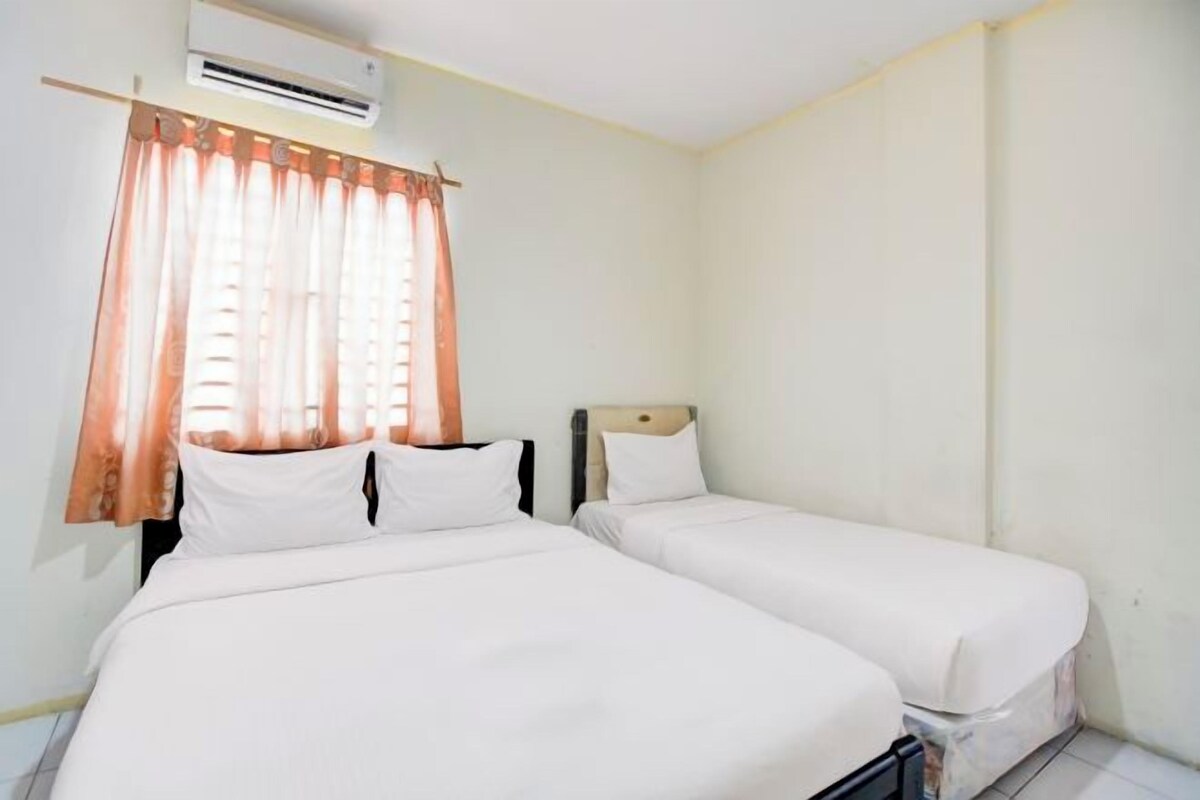 A room with two beds, a window with curtains, an air conditioning unit, and a plain wall.