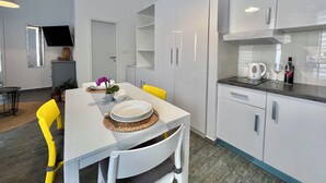 A modern kitchen with white cabinets, a dining table with yellow chairs, and a flat-screen TV mounted on the wall.