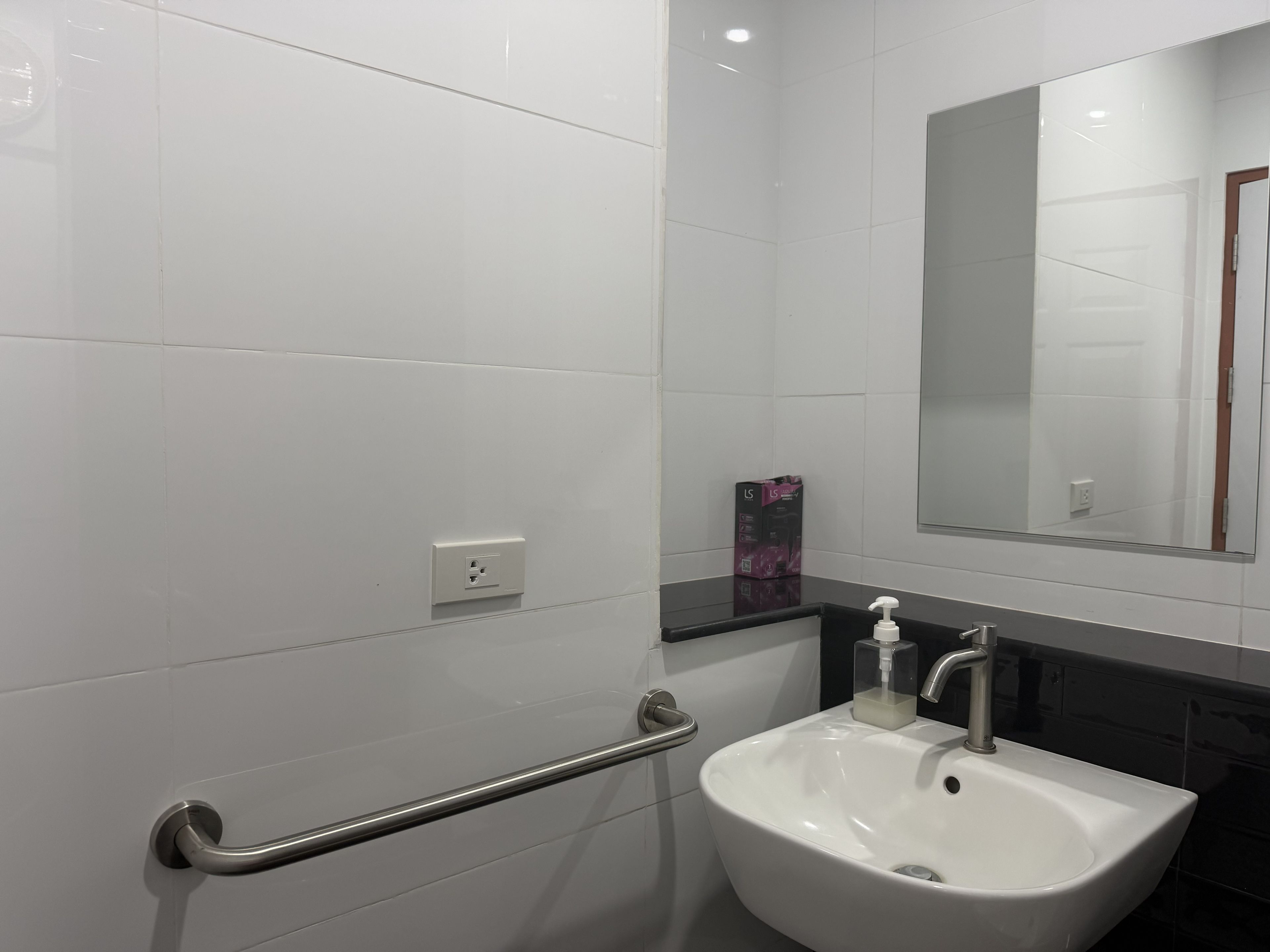 Un baño con azulejos blancos, encimera negra, lavamanos blanco, espejo y barandilla.