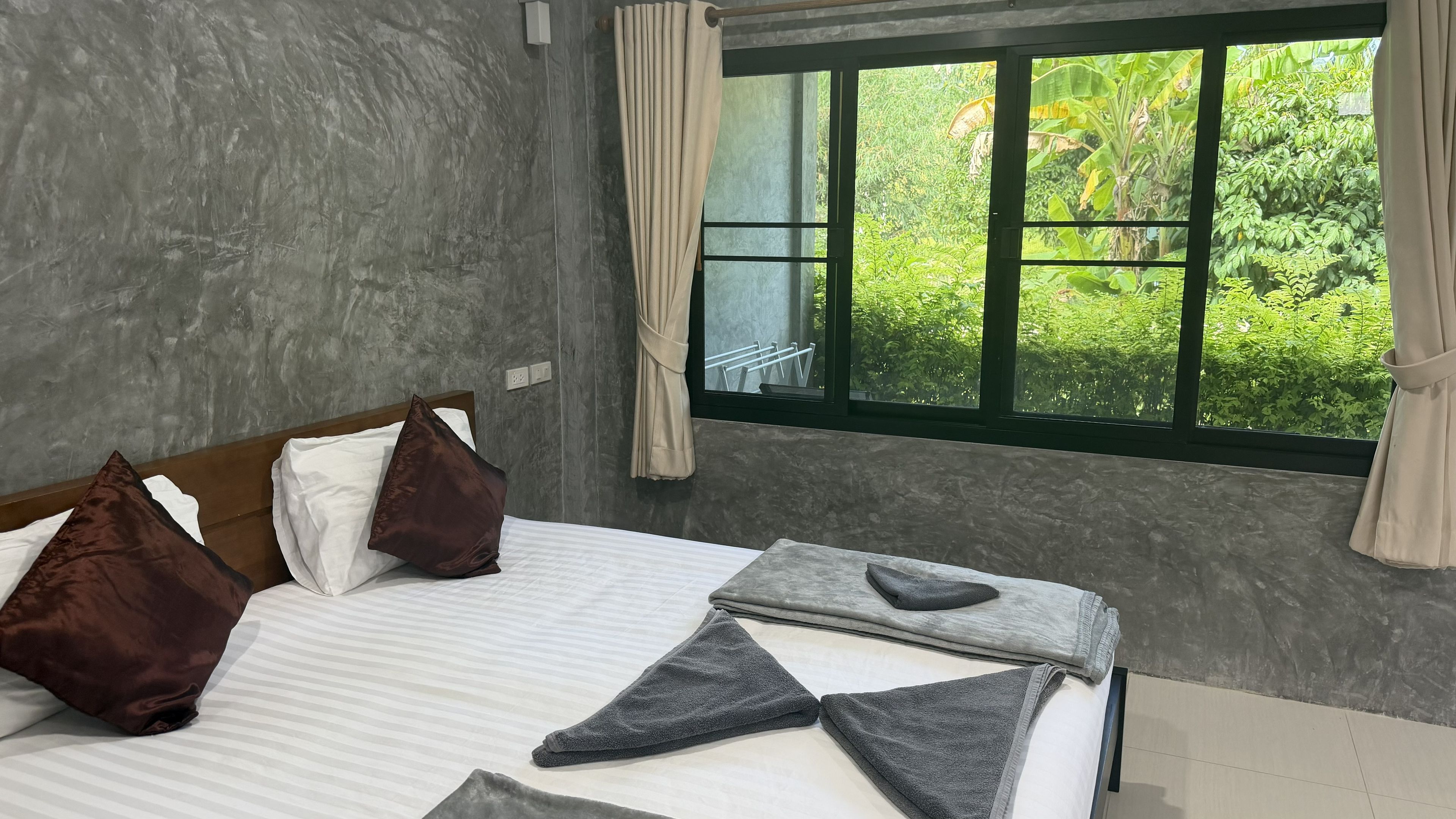 A hotel room with a bed, pillows, a window with curtains, and a view of greenery outside.