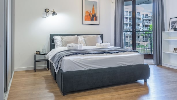 A modern bedroom with a bed, bedside tables, a cityscape artwork, and a view of a residential area through a balcony. - Apartament w Śródmieściu Odnowa (Wroclaw)