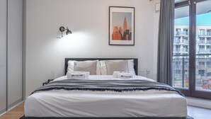 A neatly made bed with white linens and two pillows, a framed cityscape picture on the wall, and a view of a balcony with buildings in the background. - Apartament w Śródmieściu Odnowa (Wroclaw)