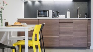 A modern kitchen with wooden cabinets, a white countertop, a microwave, a kettle, and a dining area with a white table and yellow chairs. - Apartament w Śródmieściu Odnowa (Wroclaw)