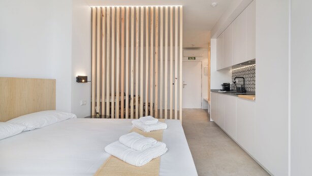 A modern hotel room with a large bed, a wooden headboard, a bedside table with a lamp, and a view into the kitchen area. - Stayaday Apartments Albufera (Madrid)