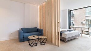 A modern hotel room with a blue sofa, two round tables, a bed, and a view of a brick building. - Stayaday Apartments Albufera (Madrid)