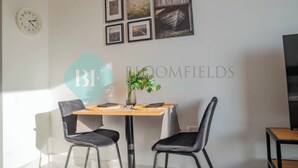 Un coin repas avec une table en bois et deux chaises, une horloge au mur et des tableaux encadrés au-dessus.