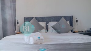 Una cama bien hecha con sábanas blancas y almohadas grises, una mesita de noche con lámpara y una lámpara de pared.