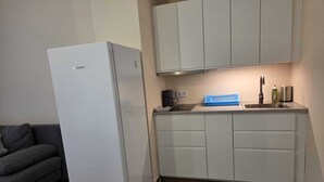 Μια μικρή, λειτουργική κουζίνα με ένα λευκό ψυγείο, έναν εντοιχισμένο νεροχύτη και έναν γκρι καναπέ.