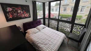Ein Schlafzimmer mit einem Bett, einem Schreibtisch, einem Stuhl und einem großen Fenster mit Blick auf Gebäude und Grünflächen.
