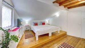 Une chambre avec un plancher en bois, un lit avec une literie blanche, un téléviseur posé sur un meuble, une table de chevet, une commode, une fenêtre avec des rideaux et une porte.