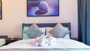 A neatly made bed with white linens and pillows, a bedside table with a lamp, and a painting on the wall. - Bloomfields Spectacular STU in Oasis (Abu Dhabi)