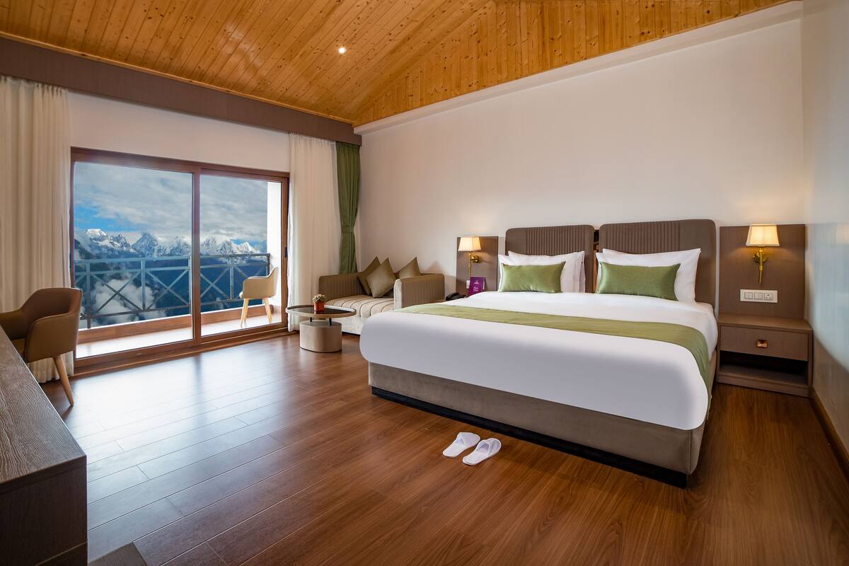 A hotel room with a large bed, a desk, and a view of mountains through a sliding glass door.