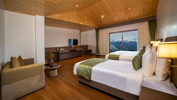 Habitación de hotel con dos camas, un sofá, un televisor, una mesita con una lámpara y vistas a montañas a través de un ventanal grande.