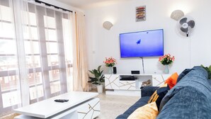 Un salon moderne doté d’un téléviseur à écran plat, d’un meuble de rangement blanc, d’un canapé bleu et d’une grande fenêtre ornée de rideaux légers.