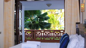 Une chambre d’hôtel avec un lit, du linge de lit blanc, une couverture bleue et une vue sur les palmiers depuis un balcon.