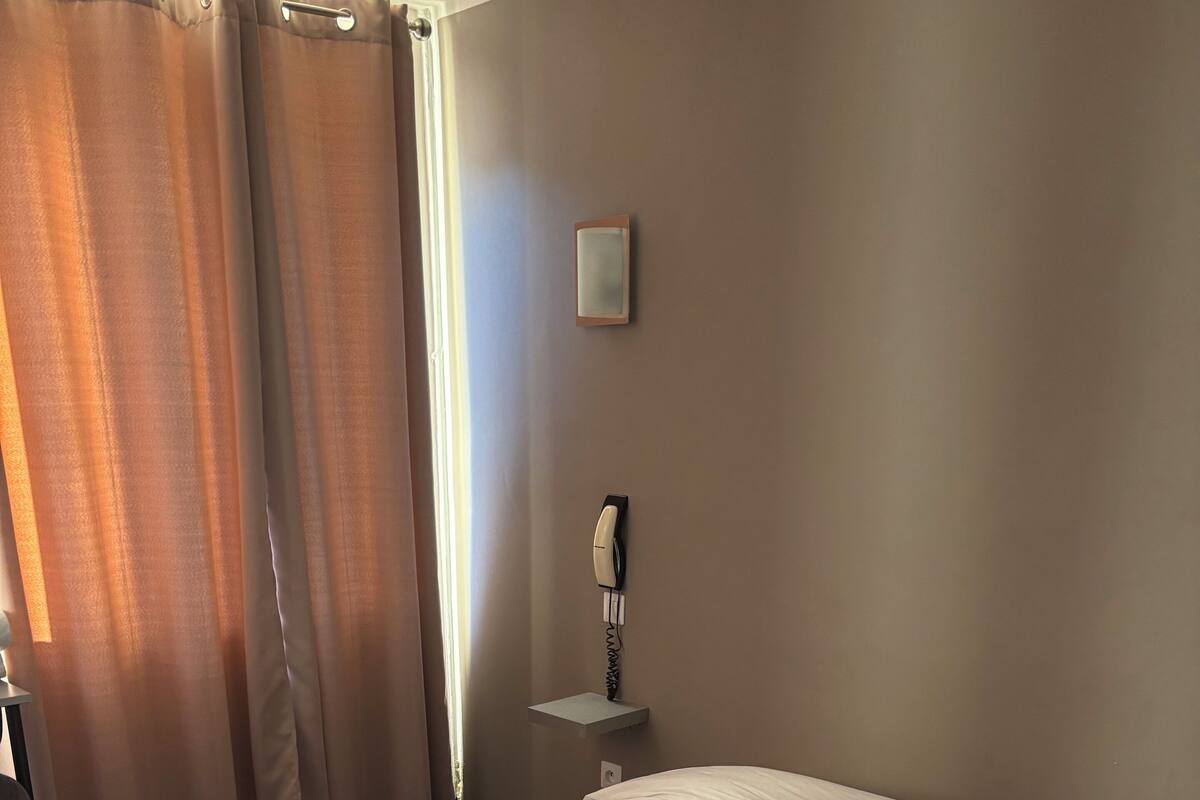 A hotel room with a bed, two pillows, a bedside table with a telephone, and a window with curtains.
