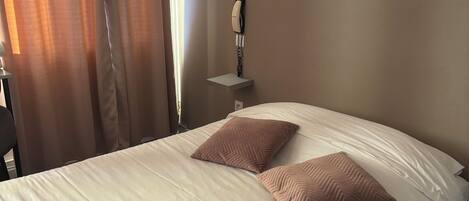 A hotel room with a bed, two pillows, a bedside table with a telephone, and a window with curtains.