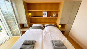 Quarto duplo com duas camas, cabeceira de madeira, uma prateleira pequena com abajur e um guarda-roupa.