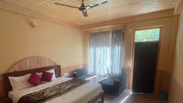 A bedroom with a wooden ceiling, a bed with white bedding and red pillows, a nightstand, a window with curtains, and a door. - Boho Riverhouse (Banjar)