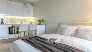 Een moderne slaapkamer met een groot bed, een nachtkastje en een kleine kitchenette op de achtergrond.