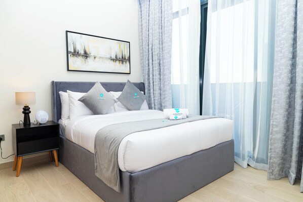 A neatly made bed with a grey headboard, white bedding, and two grey pillows. There is a bedside table with a lamp and a small decorative item. A framed abstract painting hangs above the bed.