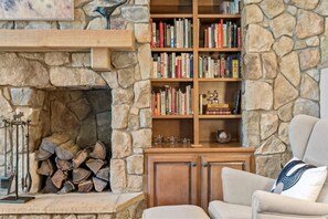 A cozy living room with a stone fireplace, a bookshelf filled with books, and a comfortable armchair. - Mountaintop Cabin Hot Tub Views Family Golf (Portola)