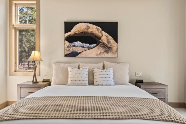 Quarto com uma cama grande, duas mesas de cabeceira, uma luminária e um quadro de uma paisagem de montanhas.