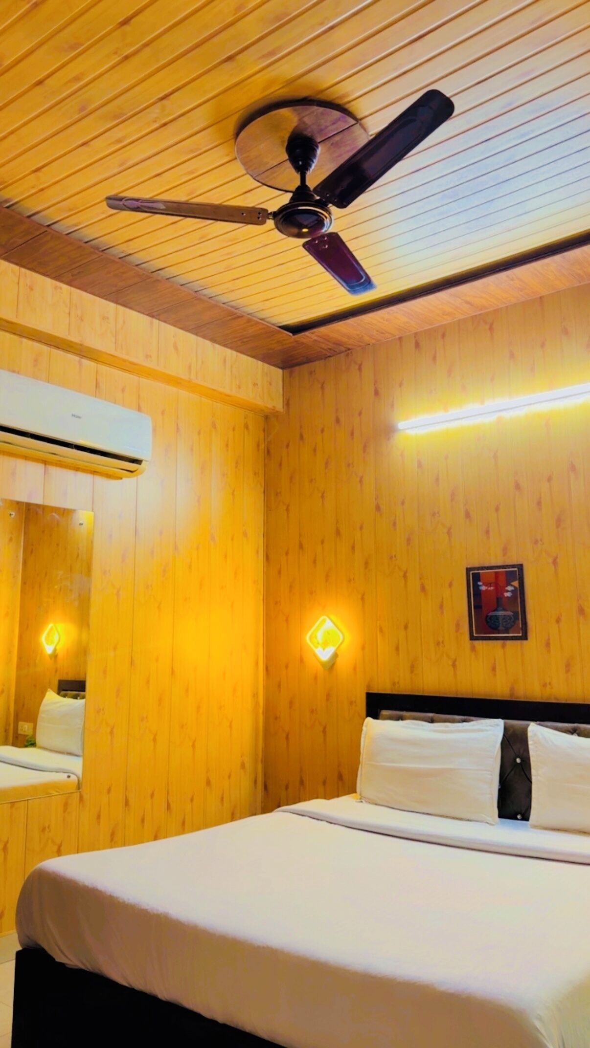 A hotel room with wooden panel walls, a ceiling fan, a bed with white bedding, a wall-mounted air conditioner, and a small framed picture.