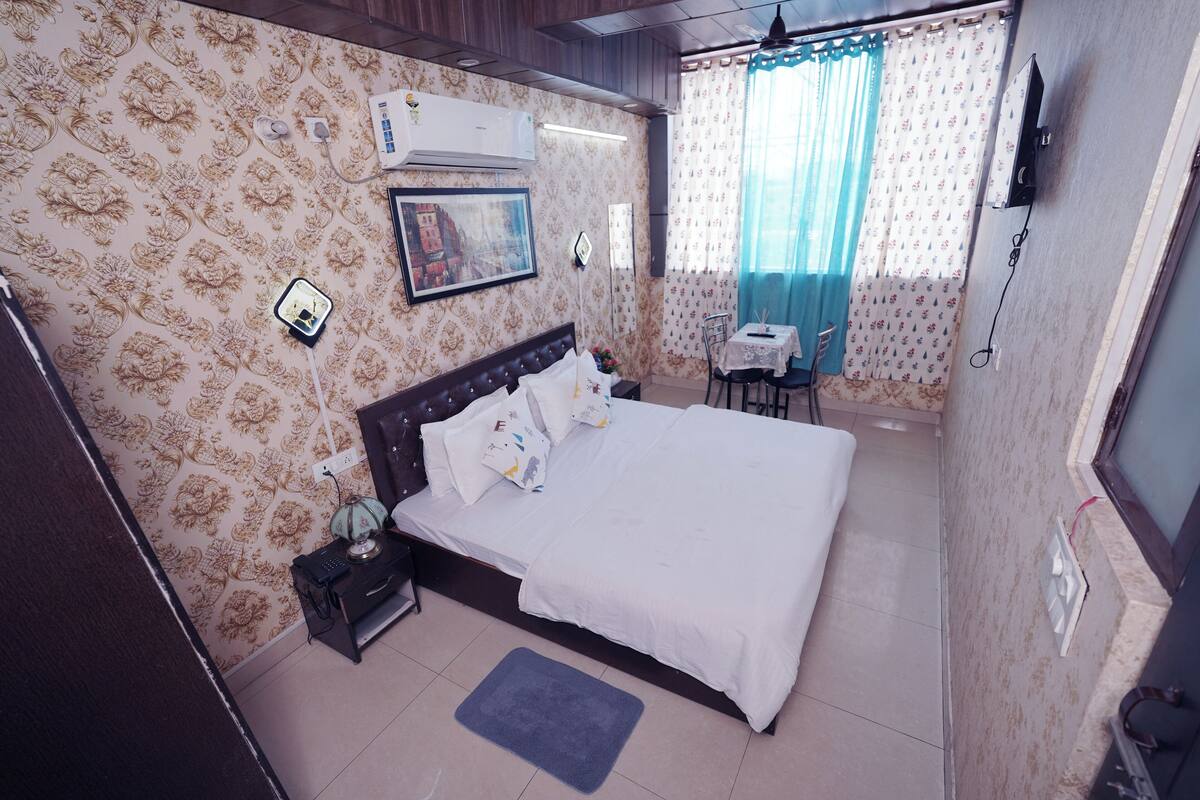 A bedroom with a bed, bedside table, nightstand, air conditioning unit, and a small desk with a chair.