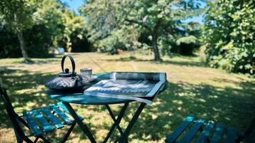 A table with a kettle, cup, and magazine in an outdoor setting with greenery.