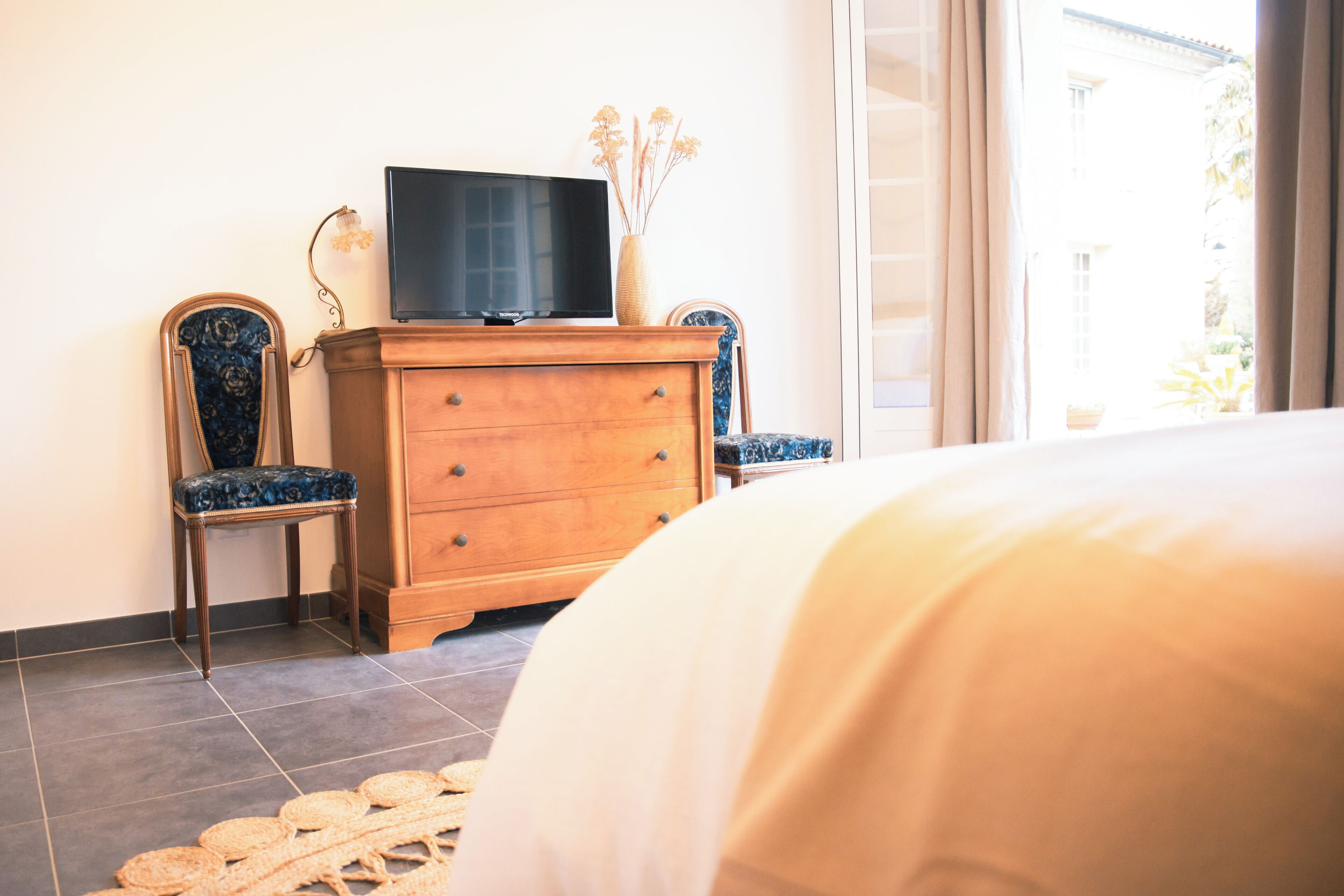 Une chambre à coucher avec un lit, une commode en bois, deux chaises, un téléviseur et une fenêtre avec des rideaux.