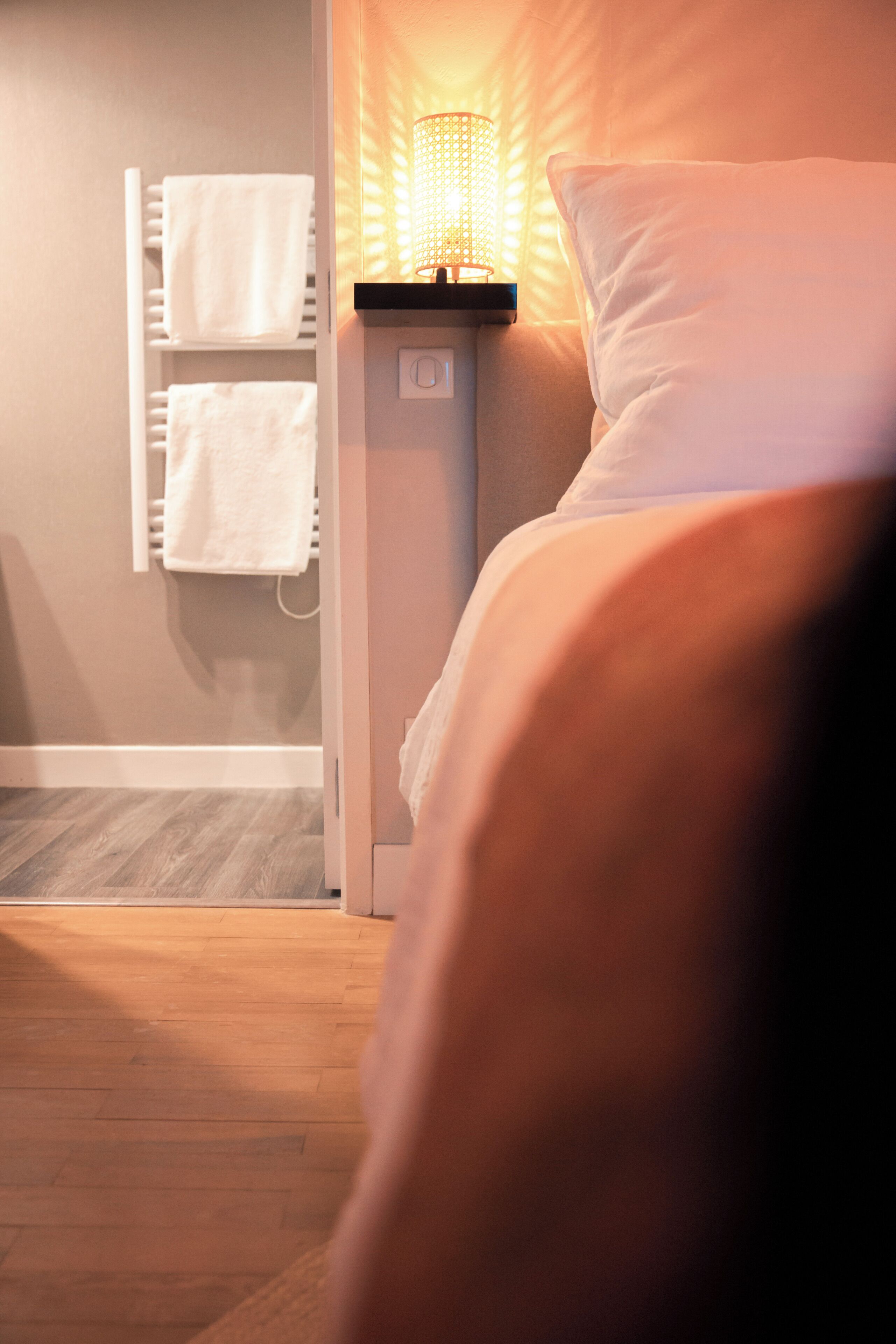 A hotel room with a bed, a bedside lamp, a towel rack, and a wooden floor.