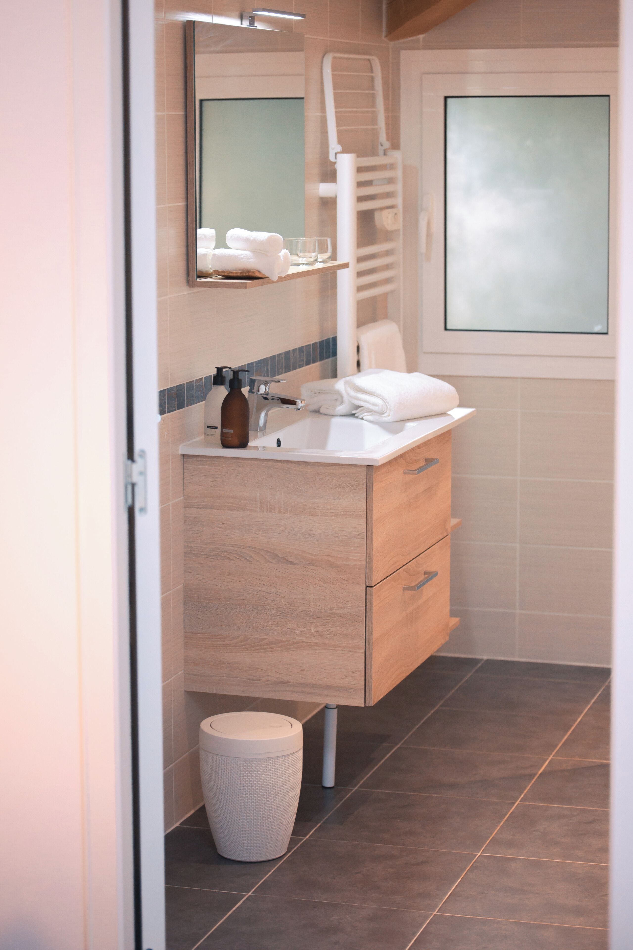 A modern bathroom with a wooden vanity, a mirror, a towel rack, and a trash bin.