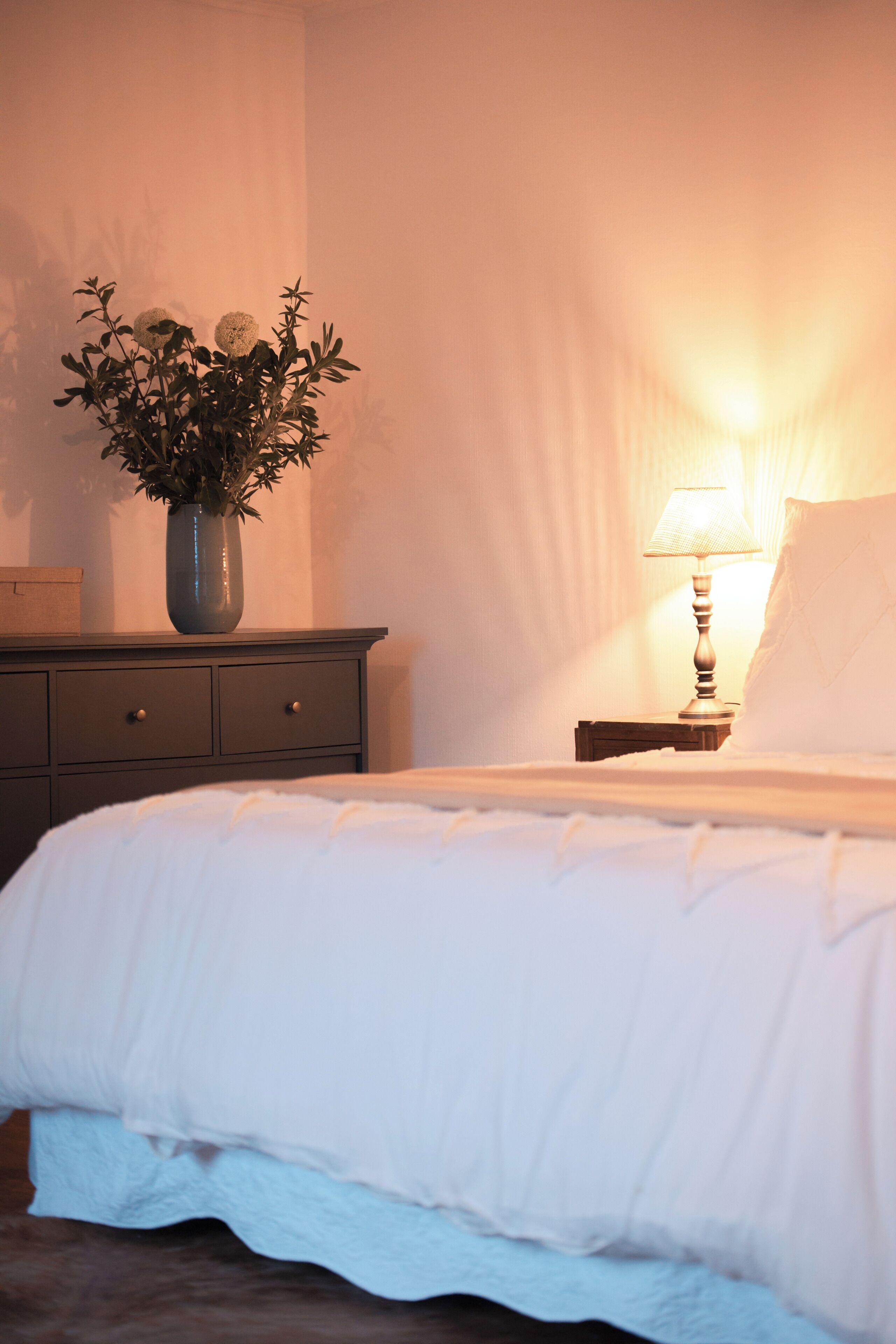 A neatly made bed with white bedding, a bedside lamp, and a vase with flowers on a dresser.