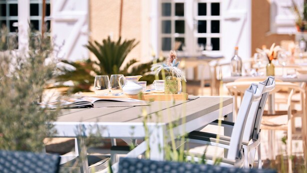 Un espace de restauration en plein air avec une table dressée pour deux personnes, entourée de chaises blanches, d’un parasol en chaume et offrant une vue sur un bâtiment doté de grandes fenêtres.