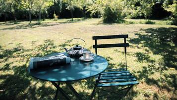 Une table ronde avec une théière, une tasse et une soucoupe, et une chaise, le tout placé dans un espace extérieur verdoyant, avec des arbres en arrière-plan.