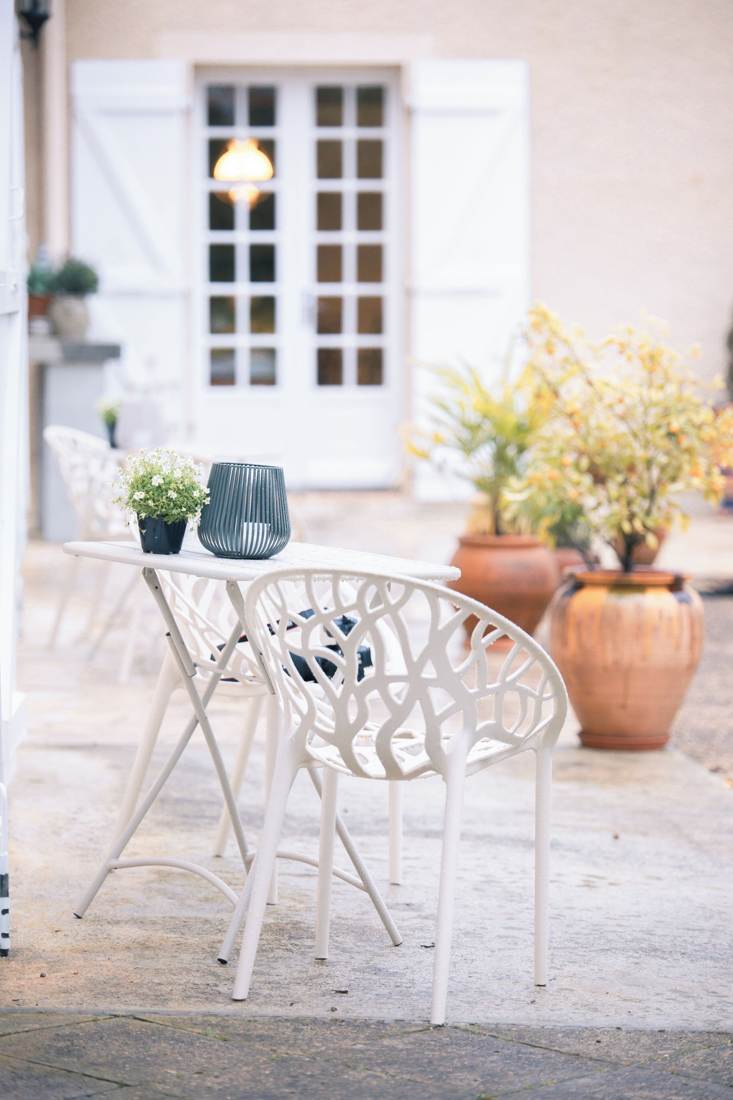 A white outdoor seating area with a table and chairs, potted plants, and a building with a window and door in the background.