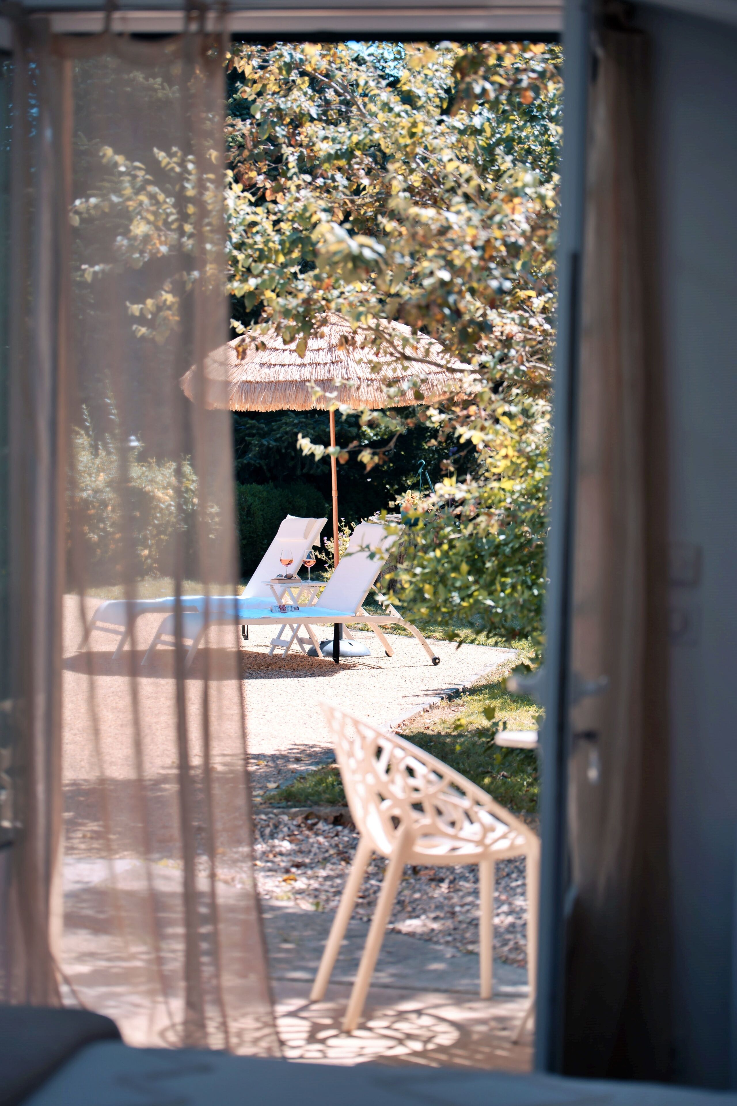 Une vue à travers une fenêtre sur un espace piscine, avec des chaises longues et un parasol en chaume.
